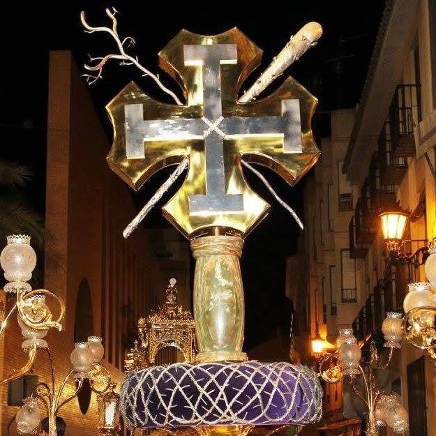 El trono-insignia de la cofradía: la cruz flordelisada en oro con la cruz potenzada superpuesta y los flagelos en plata, alzada de noche durante la procesión.